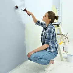 Kobieta w kratę maluje ścianę na jasny kolor, pokazując, jak pomalować pokój. W tle drabina i wiaderka z farbą.