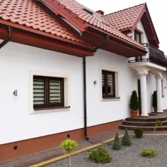 Biały dom z brązowym cokołem wokół, czerwonym dachem i ozdobnymi schodami.