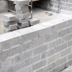 Budowa muru z szarego bloczka betonowego. Widać stosy bloczków, przygotowane do murowania.