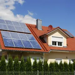Dach domu pokryty czerwonymi dachówkami, na którym zamontowano panele fotowoltaiczne. Widać różne rodzaje paneli fotowoltaicznych, które zbierają energię słoneczną.