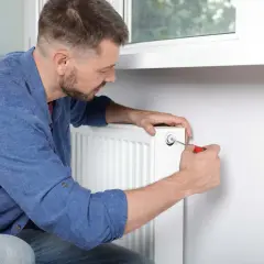Mężczyzna odpowietrza grzejnik, używając śrubokręta. Zastanawia się, od którego grzejnika zacząć odpowietrzanie.