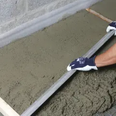 Ręce w rękawiczkach wygładzają świeży beton, który leży na siatce zbrojeniowej i styropianie. To praca przy budowie, gdzie styrobeton czy styropian odgrywają kluczową rolę.