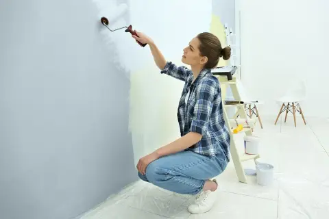 Kobieta w kratę maluje ścianę na jasny kolor, pokazując, jak pomalować pokój. W tle drabina i wiaderka z farbą.