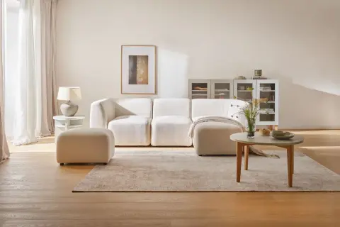 Jasny salon: biel, szary i drewno. Miękka sofa, puf, stolik kawowy i dywan tworzą przytulną przestrzeń.