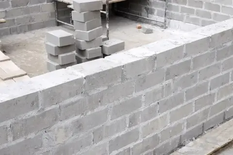 Budowa muru z szarego bloczka betonowego. Widać stosy bloczków, przygotowane do murowania.
