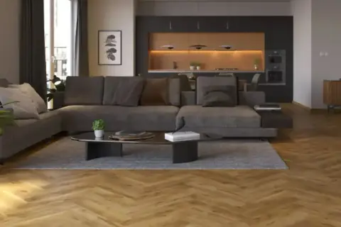 Nowoczesny salon z szarą sofą i ciemną podłogą. W tle kuchnia z drewnianymi frontami i oświetleniem.