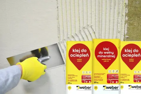 Gąbkowanie kleju na elewacji. Ręka w żółtej rękawicy rozprowadza klej na izolacji, obok worki z klejem do ociepleń.