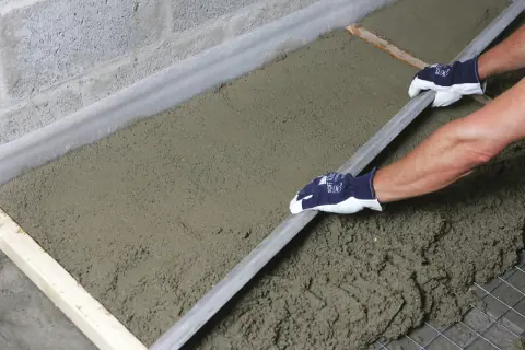 Ręce w rękawiczkach wygładzają świeży beton, który leży na siatce zbrojeniowej i styropianie. To praca przy budowie, gdzie styrobeton czy styropian odgrywają kluczową rolę.