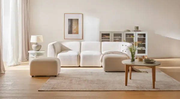 Jasny salon: biel, szary i drewno. Miękka sofa, puf, stolik kawowy i dywan tworzą przytulną przestrzeń.