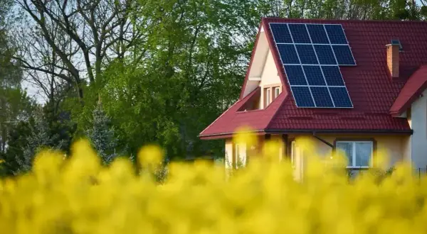 Dom z panelami fotowoltaicznymi na dachu, otoczony kwitnącym rzepakiem. Rozbudowa instalacji fotowoltaicznej w malowniczym otoczeniu.