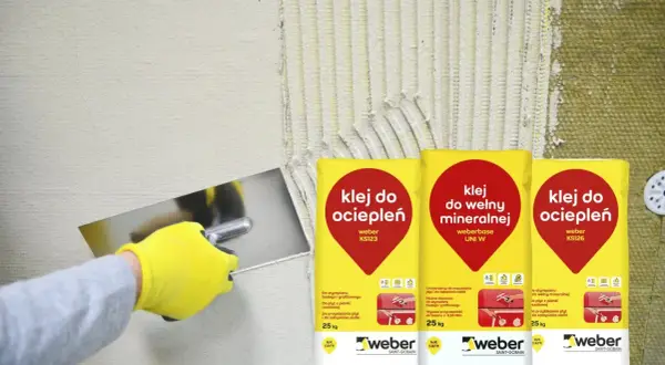 Gąbkowanie kleju na elewacji. Ręka w żółtej rękawicy rozprowadza klej na izolacji, obok worki z klejem do ociepleń.