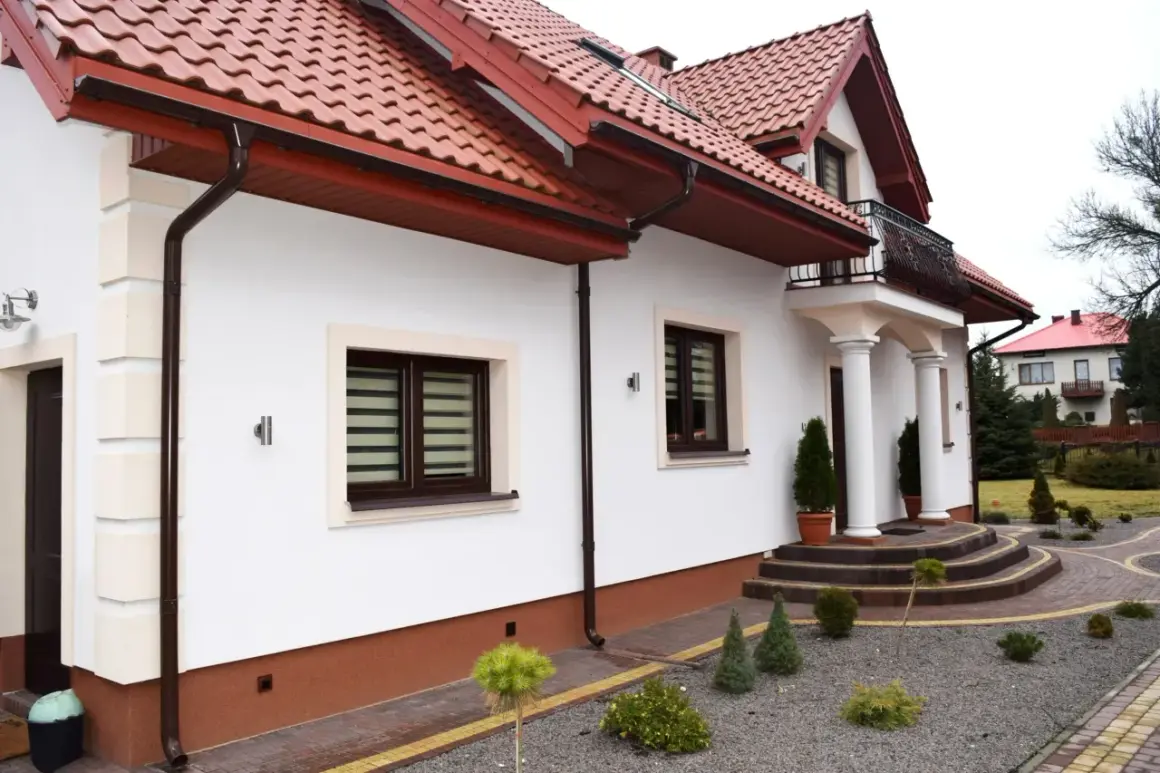 Biały dom z brązowym cokołem wokół, czerwonym dachem i ozdobnymi schodami.