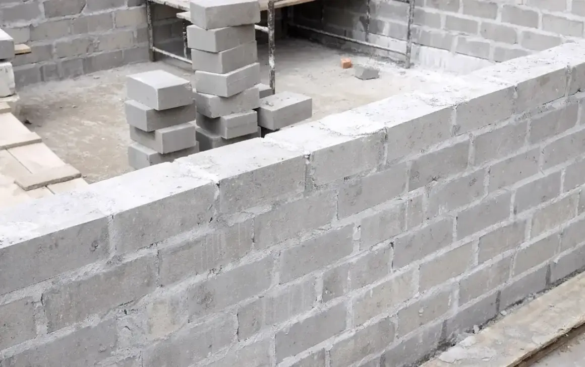 Budowa muru z szarego bloczka betonowego. Widać stosy bloczków, przygotowane do murowania.