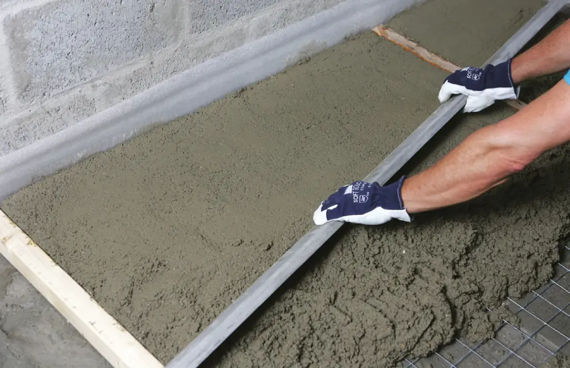 Ręce w rękawiczkach wygładzają świeży beton, który leży na siatce zbrojeniowej i styropianie. To praca przy budowie, gdzie styrobeton czy styropian odgrywają kluczową rolę.