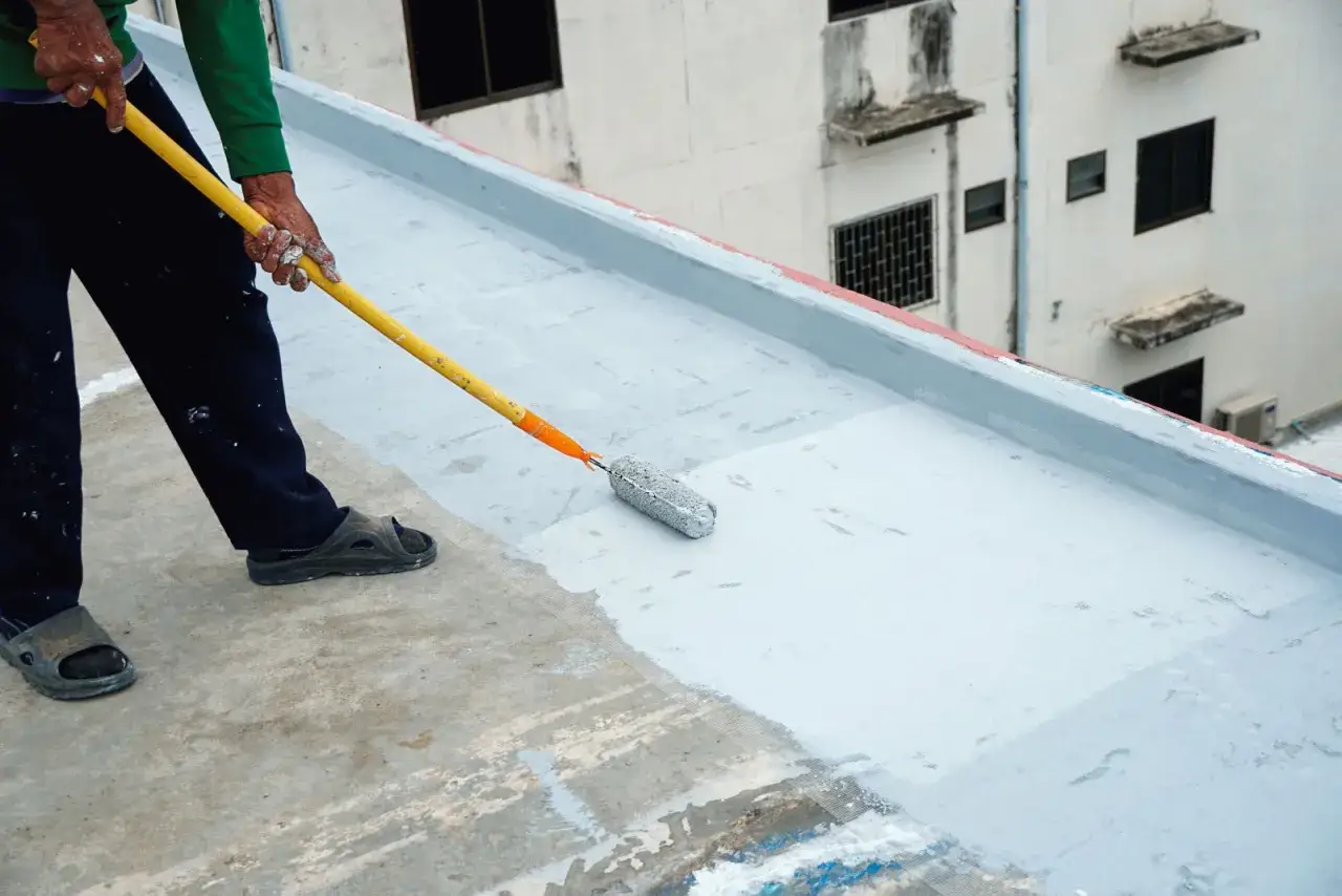 Pracownik nakłada wałkiem szarą folia w płynie na zewnątrz, zabezpieczając dach przed wilgocią.