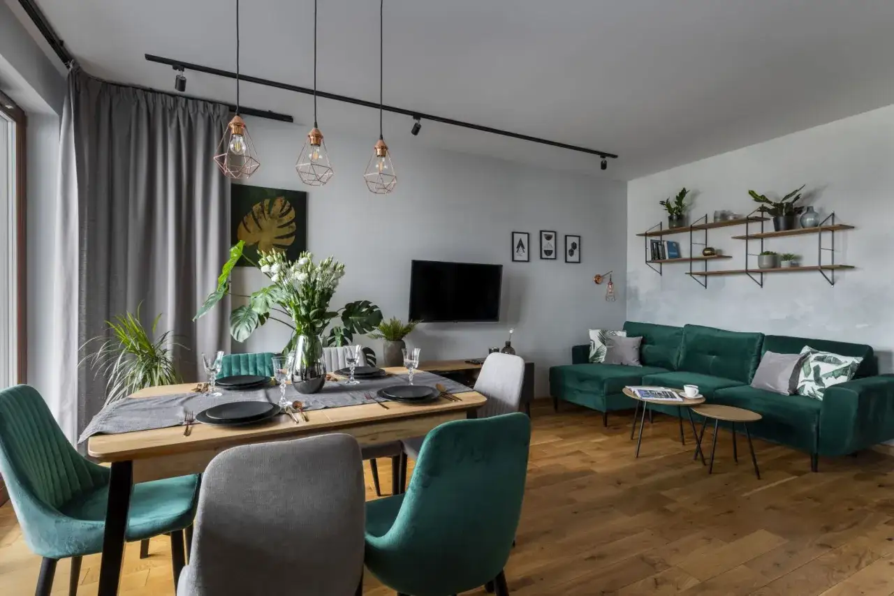 Elegancka jadalnia z zielonymi akcentami. Kolor cappuccino z czym łączyć? Szarość zasłon i krzeseł tworzy przytulną bazę dla butelkowej zieleni sofy i dodatków.
