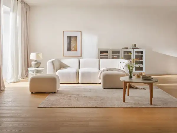Jasny salon: biel, szary i drewno. Miękka sofa, puf, stolik kawowy i dywan tworzą przytulną przestrzeń.