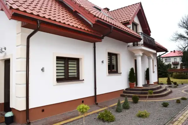 Biały dom z brązowym cokołem wokół, czerwonym dachem i ozdobnymi schodami.